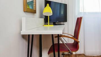 Quarto com escrivaninha branca, abajur amarelo, cadeira vermelha, televisão preta e janela com cortinas brancas.