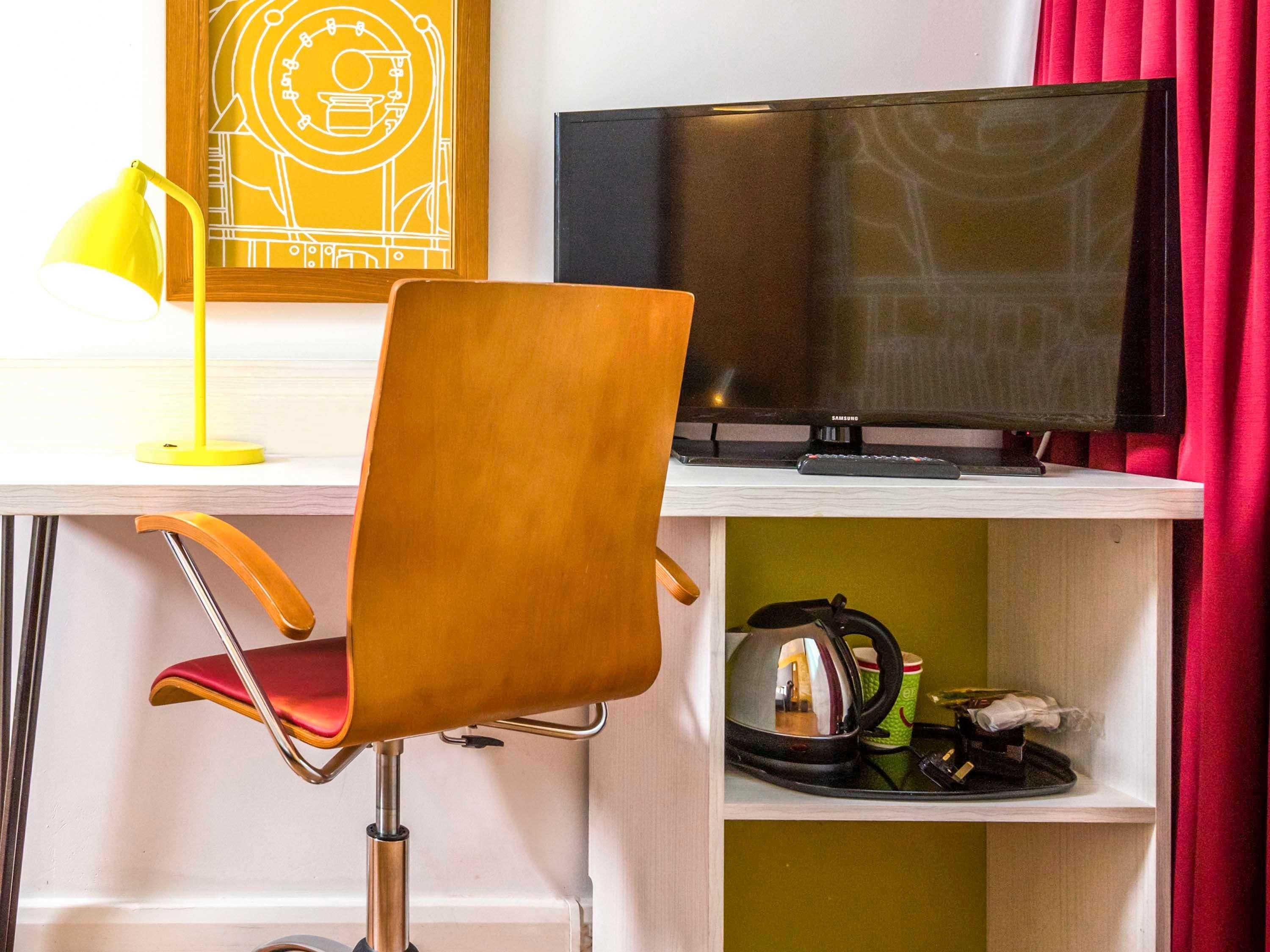 Et skrivebord med en computer, en stol, en lampe og en hylde med elkedel og kop.
