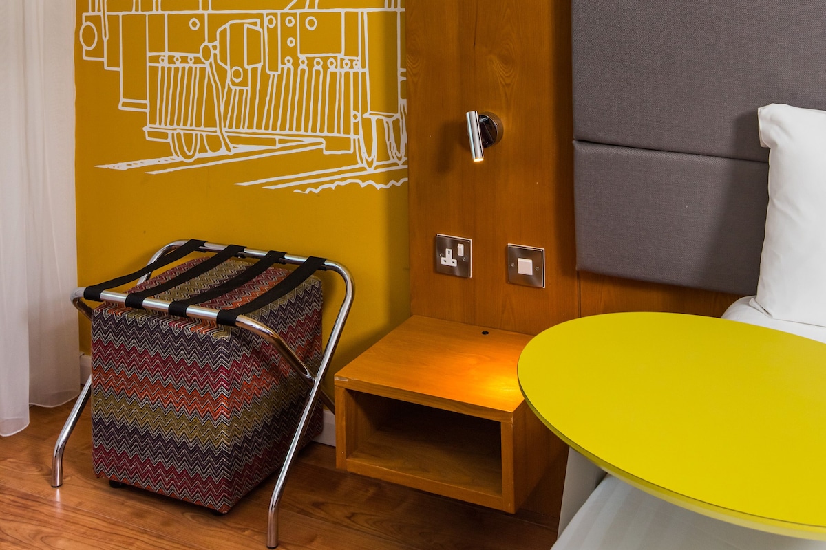 A room with a yellow wall, a wooden door, a gray upholstered headboard, a small wooden table, and a colorful chair.