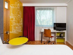 A hotel room with a bed, a desk with a TV, a chair, a yellow table, a red curtain, and a window with a view. - ibis Styles Crewe (Crewe)