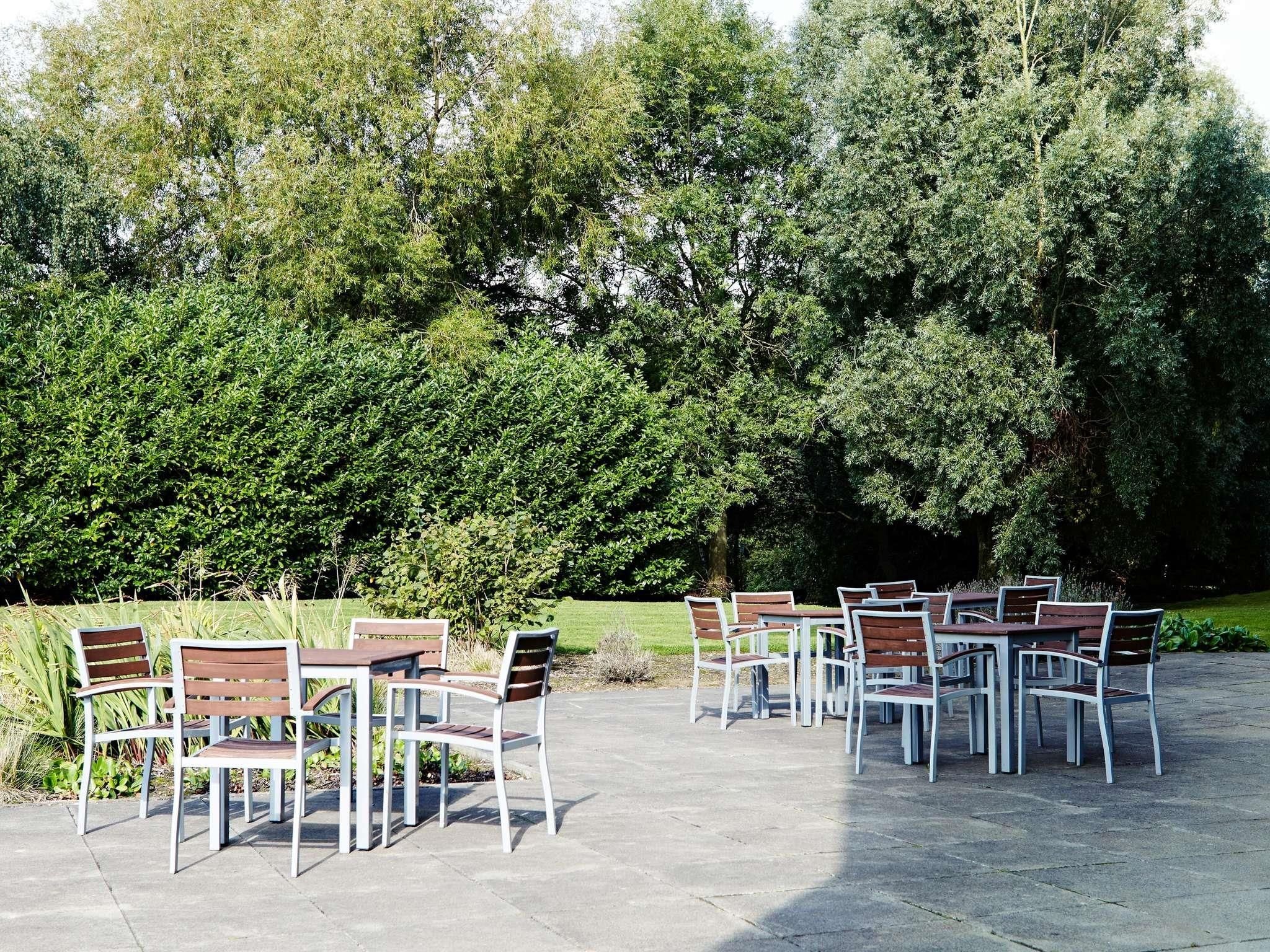 An outdoor seating area with tables and chairs arranged on a paved surface, surrounded by dense greenery and trees.