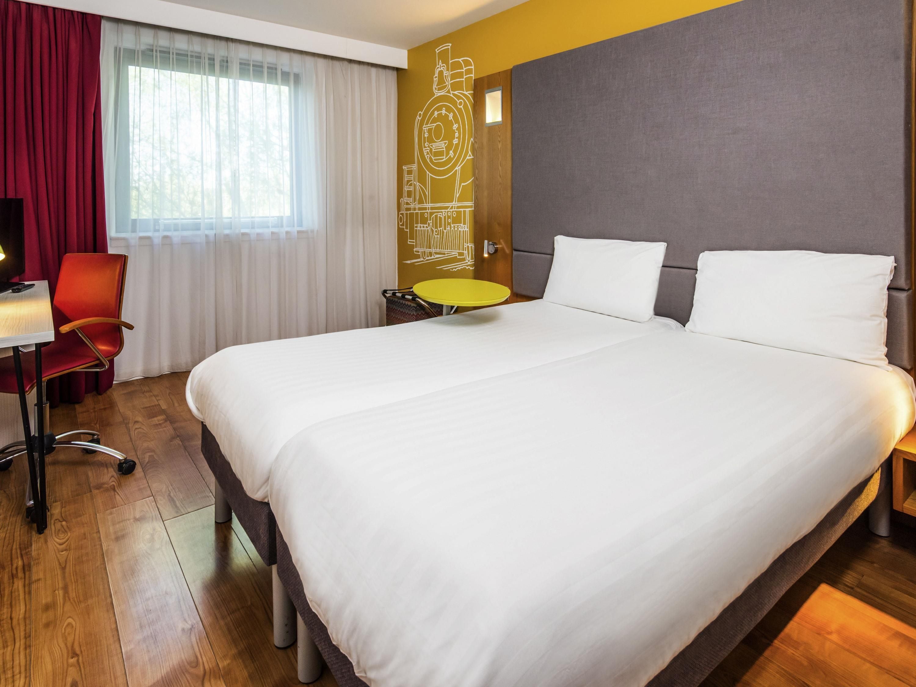 Hotelkamer met een groot bed, een bureau met stoel, een klein rond tafeltje en een raam met gordijnen.