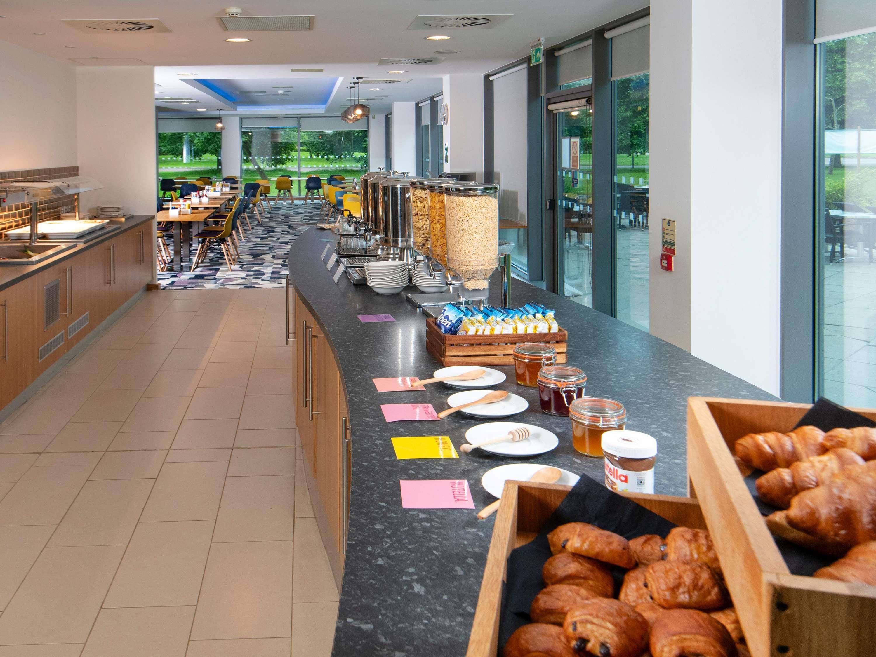 A buffet area with a variety of food items, including pastries and condiments, and a view of a park through large windows.