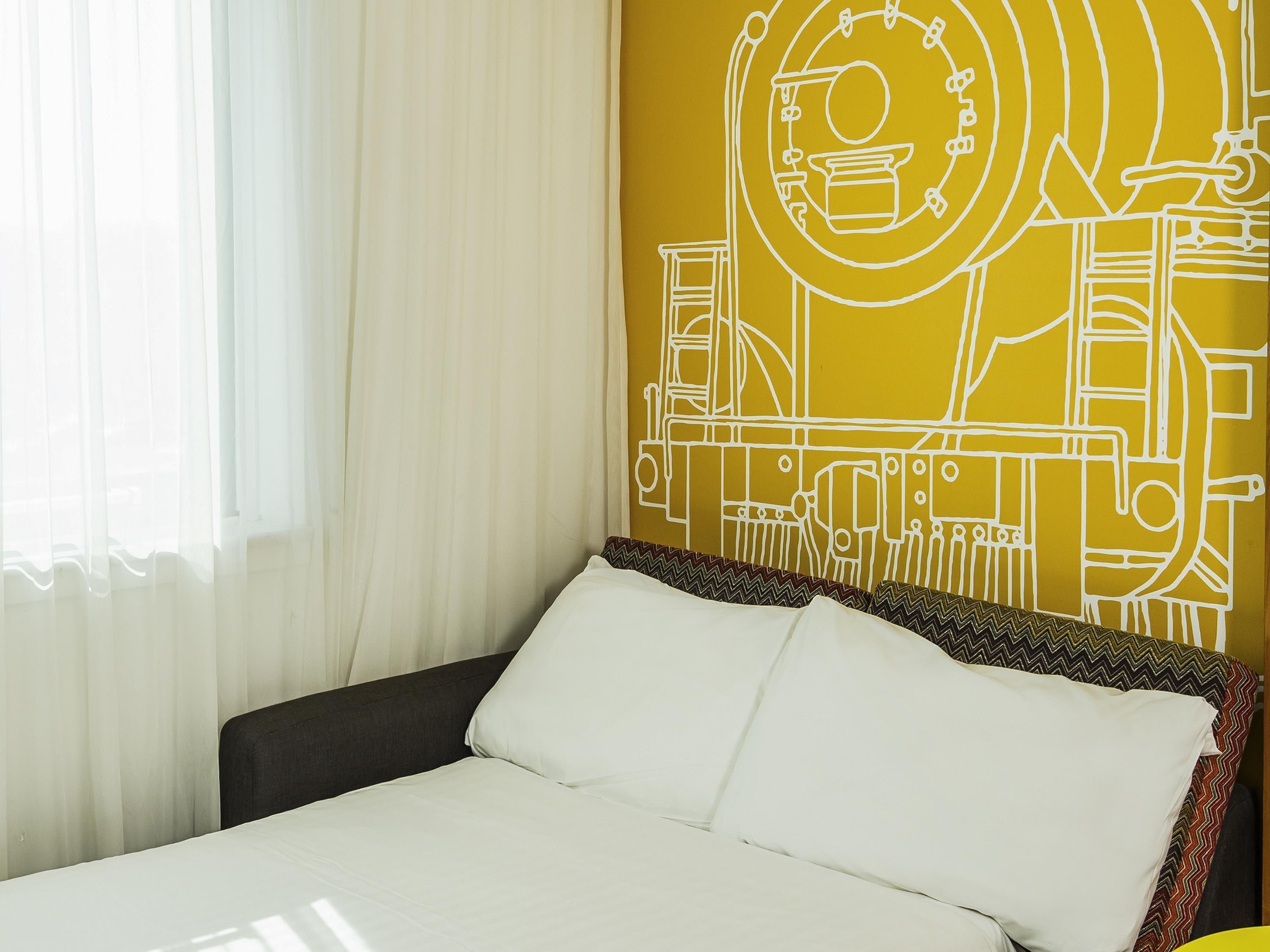 A hotel room with a bed, white curtains, and a wall with a yellow abstract design.