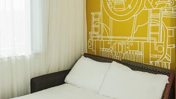A hotel room with a bed, white curtains, and a wall with a yellow abstract design.