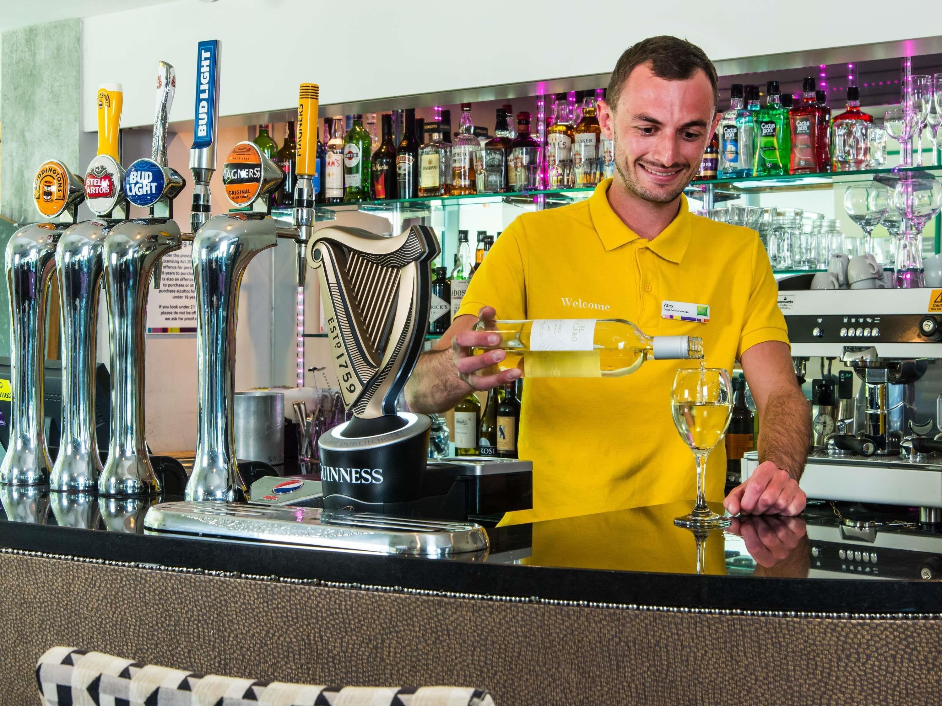 A bartender in a yellow shirt pouring a drink from a Bud Light tap into a glass. Behind him are multiple beer taps, including Guinness and Stella Artois.