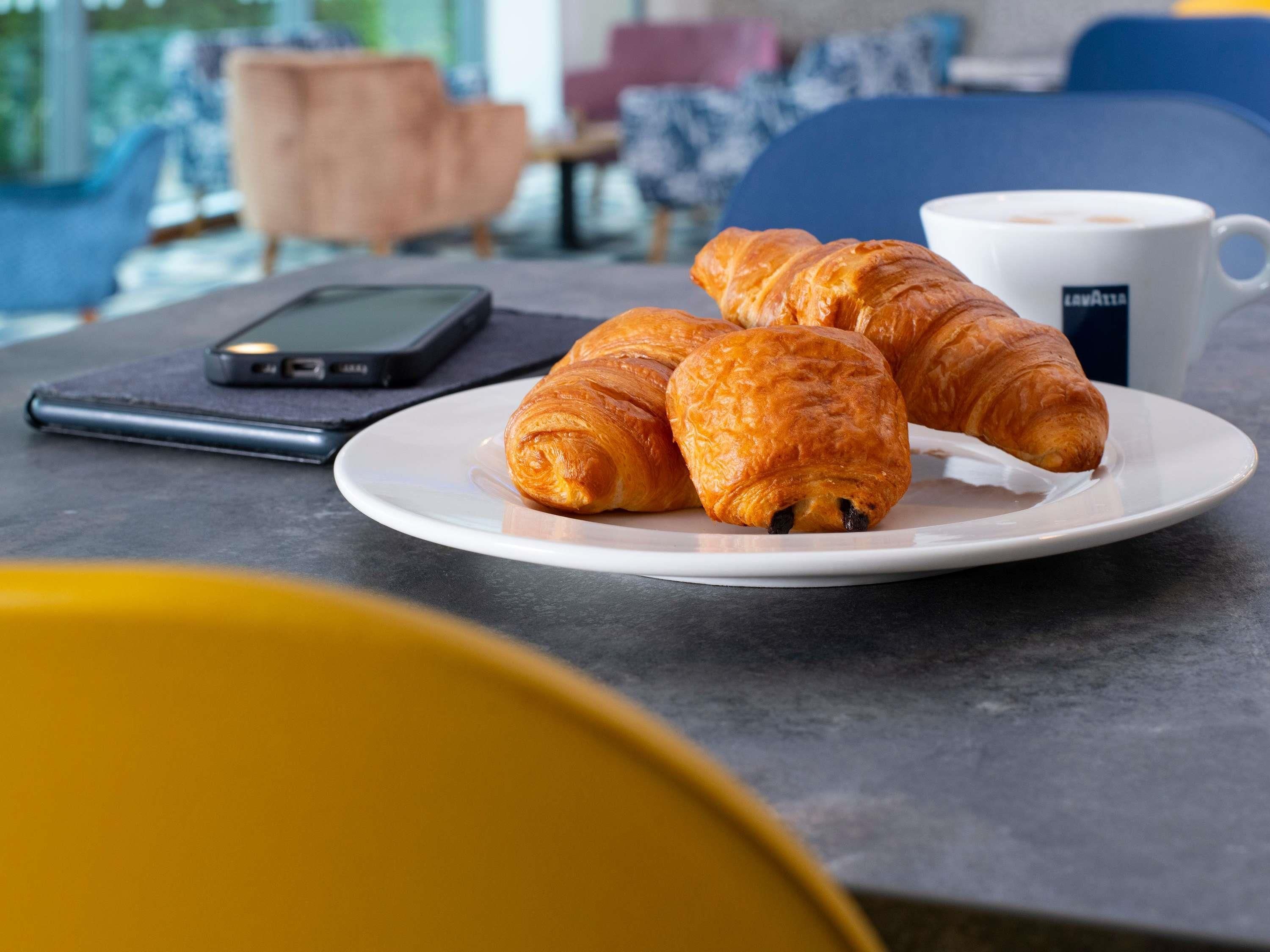 Ein Teller mit Croissants und einer Kaffeetasse auf einem Tisch, im Vordergrund ein Smartphone und ein Stuhl.