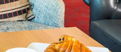 A croissant on a white plate with a cup, placed on a wooden table in a room with a red carpet and a blue leather couch.