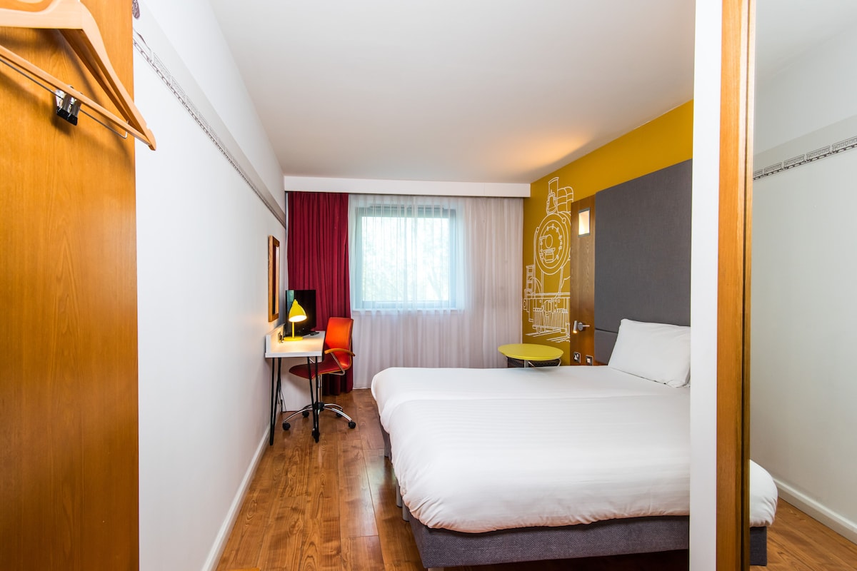 A hotel room with a bed, a desk with a lamp, a chair, a window with curtains, and a door.