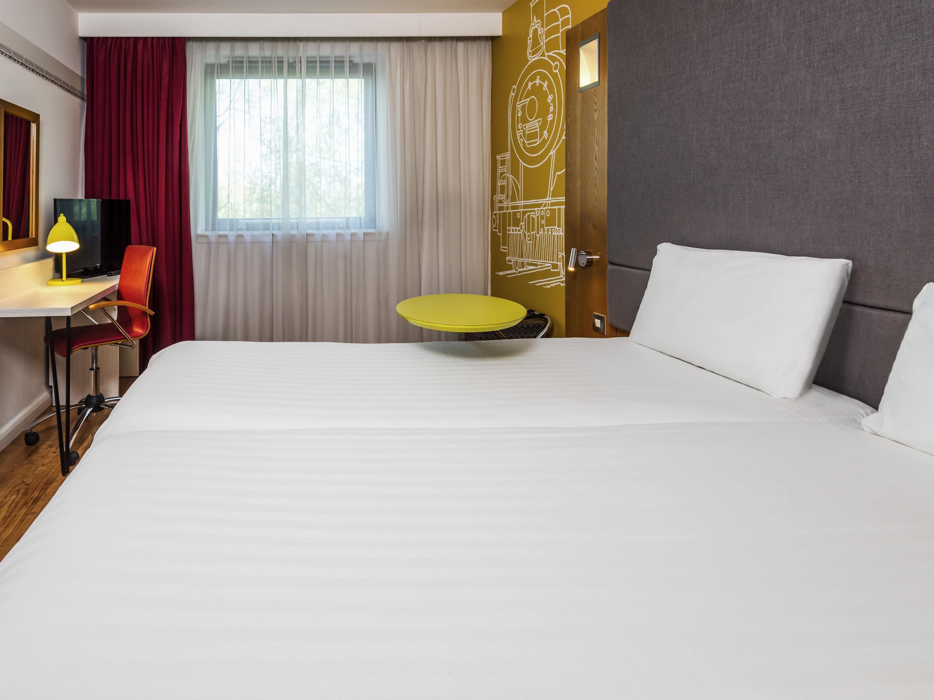 A hotel room with a large bed, a desk with a chair, a small round table, and a window with curtains.