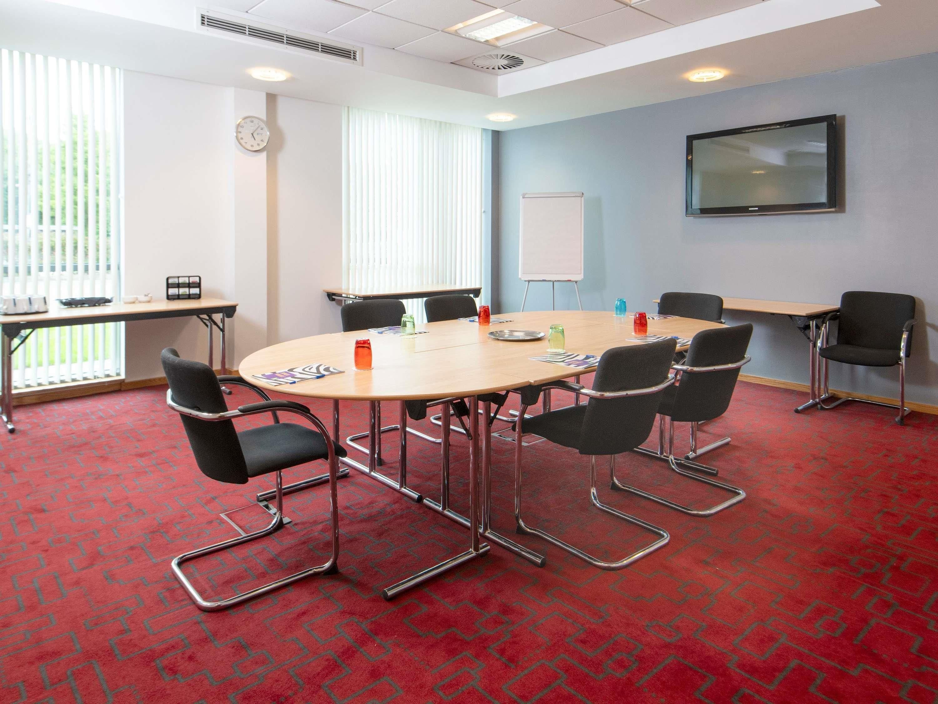 Une salle de conférence avec une table ronde, des chaises noires, un tableau blanc et un téléviseur fixé au mur.