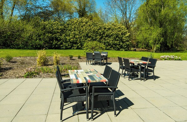 An outdoor patio with tables and chairs, surrounded by a well-maintained lawn and trees.