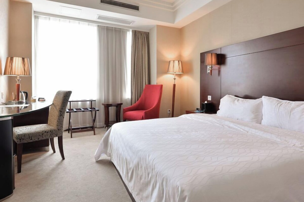 A hotel room with a large bed, a desk with a chair, a red chair, a lamp, and a window with curtains.
