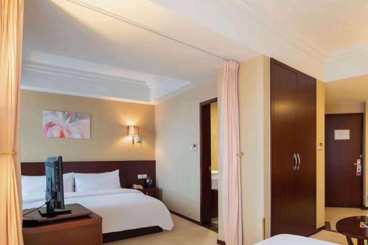 A hotel room with a bed, bedside table, lamp, artwork, and a wardrobe.
