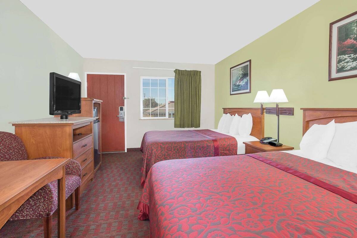 A hotel room with two beds, a desk, a TV, a window with curtains, and a painting on the wall.