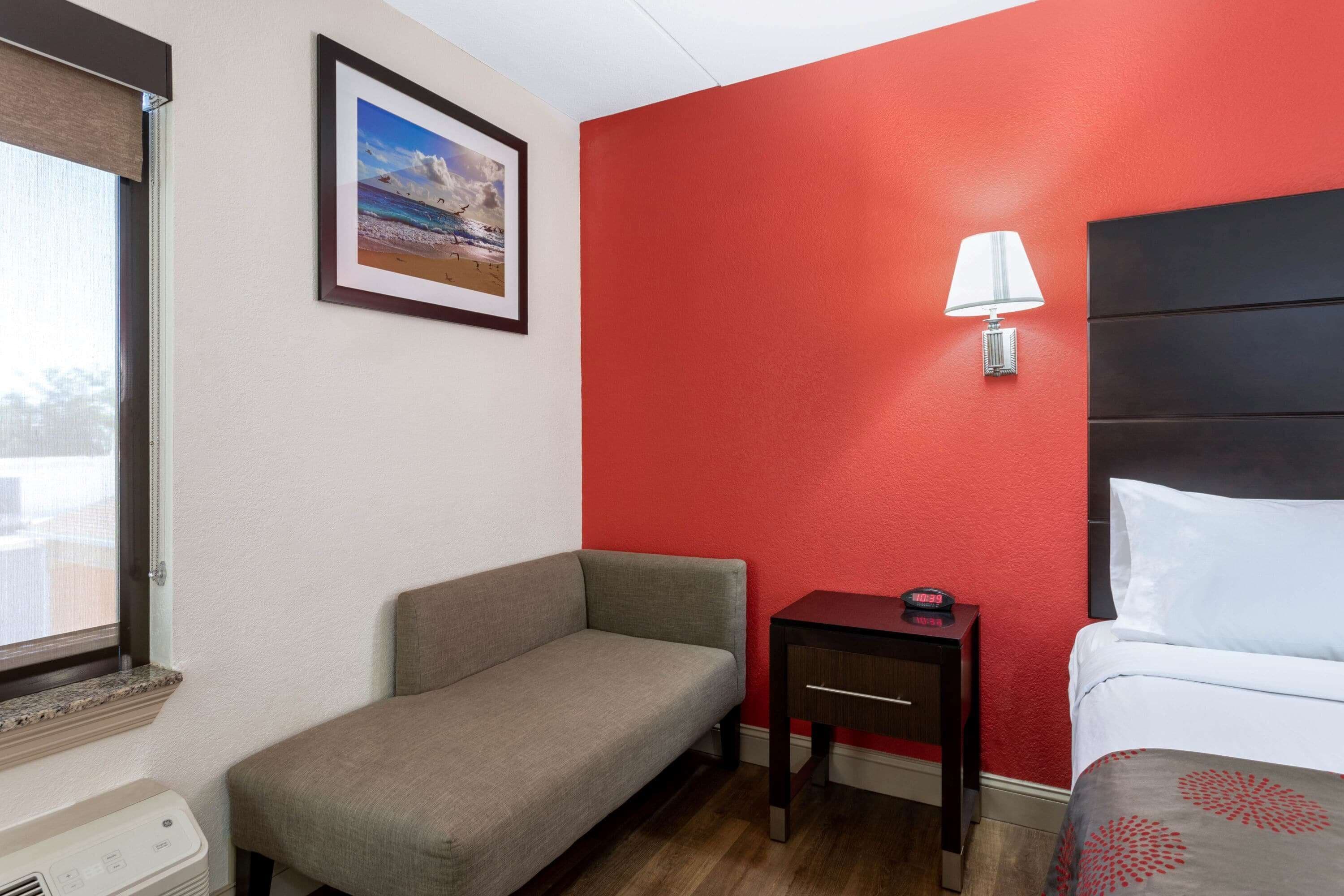 A hotel room with a red accent wall, a grey sofa, a small bedside table with a lamp, and a bed with white bedding.