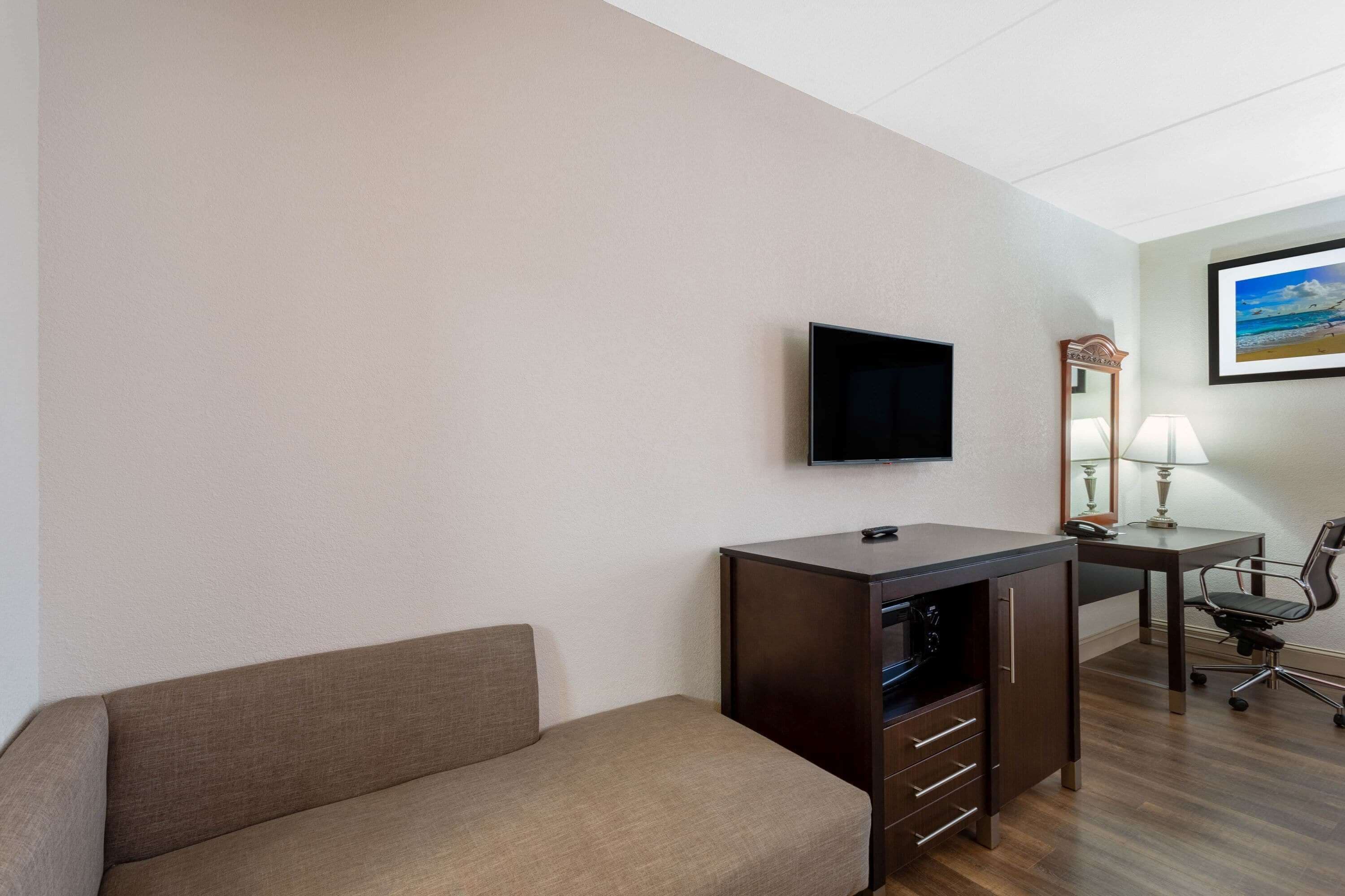 A hotel room with a sofa, a desk with a computer, a TV, a lamp, and a chair.