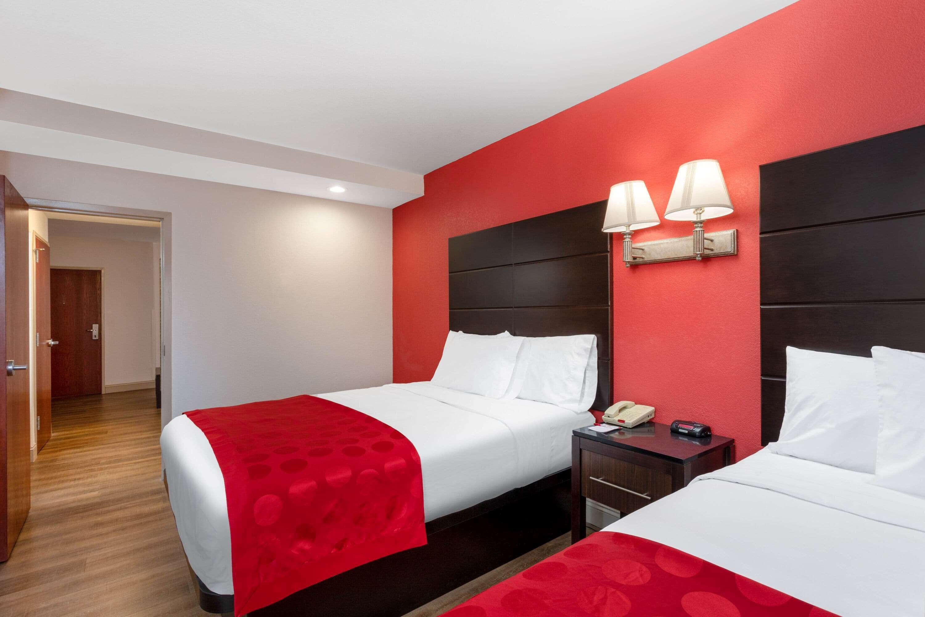 Habitación de hotel con dos camas, una pared de acento roja y una mesita de noche con teléfono y control remoto.