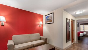 Habitación de hotel con una pared de acento roja, un sofá color beige, una mesa de centro de madera, dos lámparas de noche, un cuadro enmarcado y un acceso a un dormitorio con dos camas.