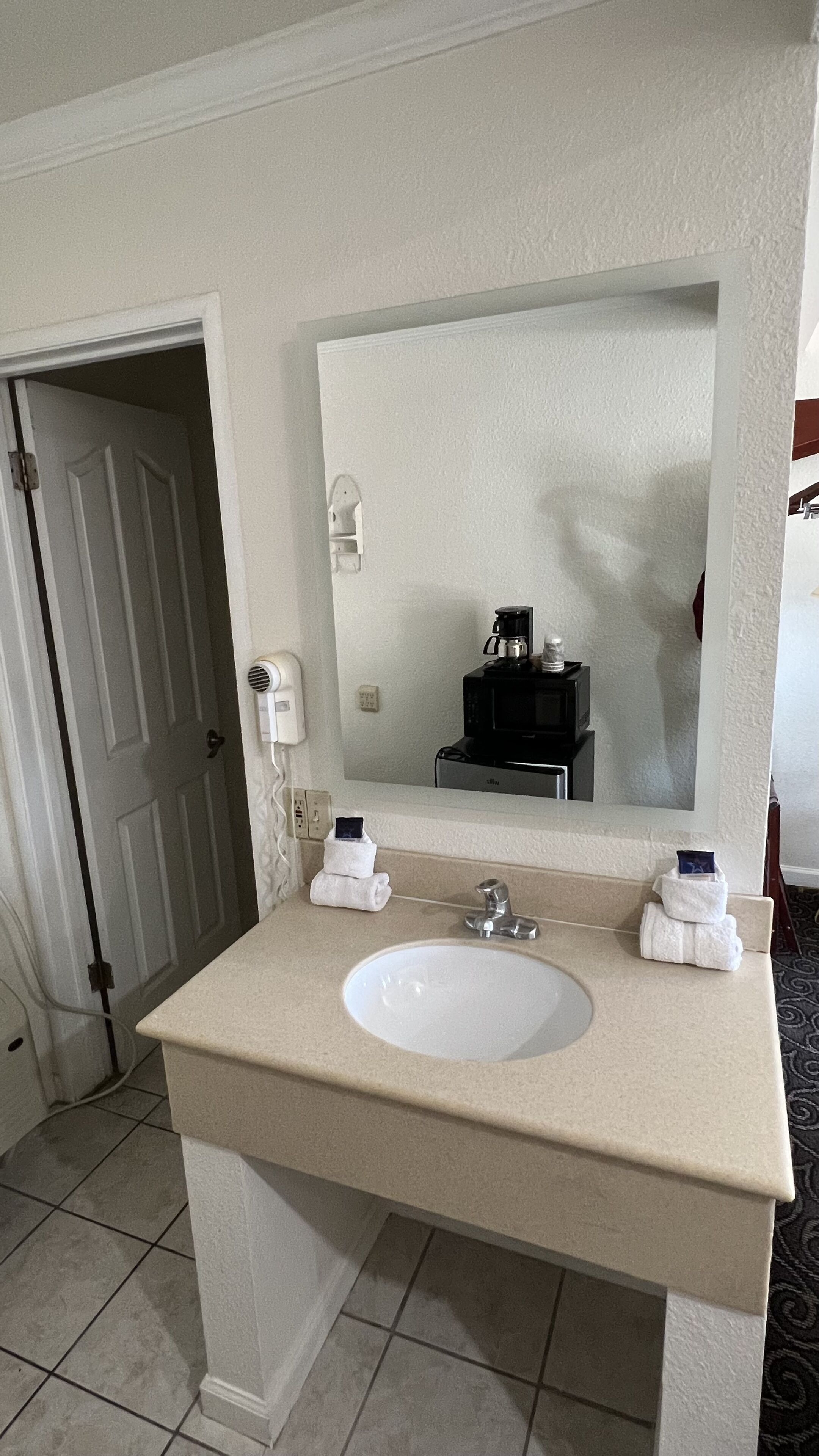 Un baño con lavamanos blanco, un espejo, un perchero para toallas y una cafetera.