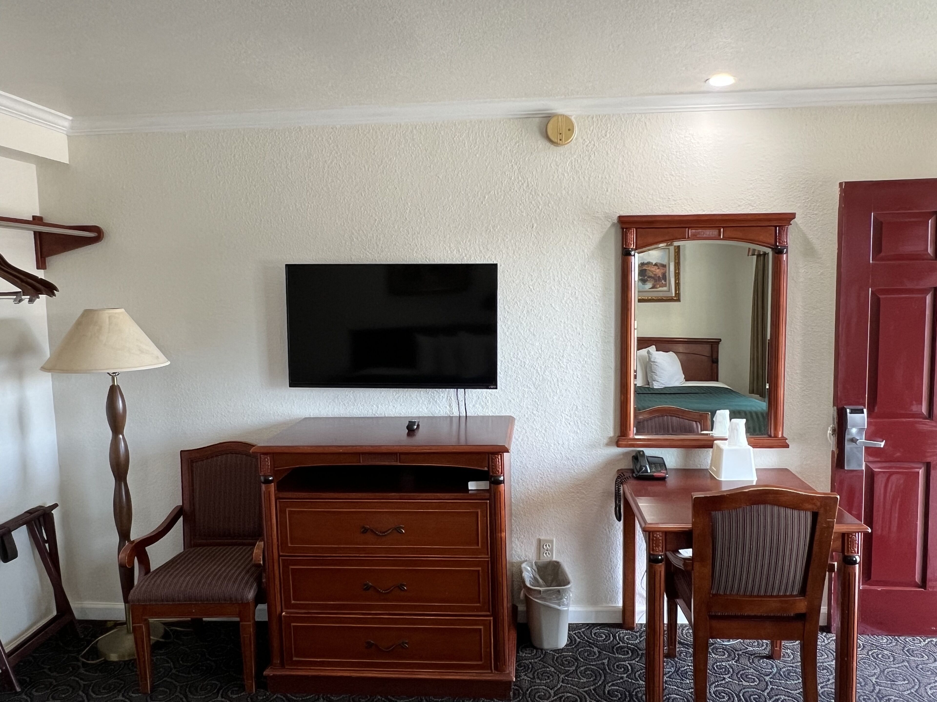 A hotel room with a flat-screen TV mounted on the wall, a wooden dresser with drawers, a chair, a desk with a telephone, and a mirror.