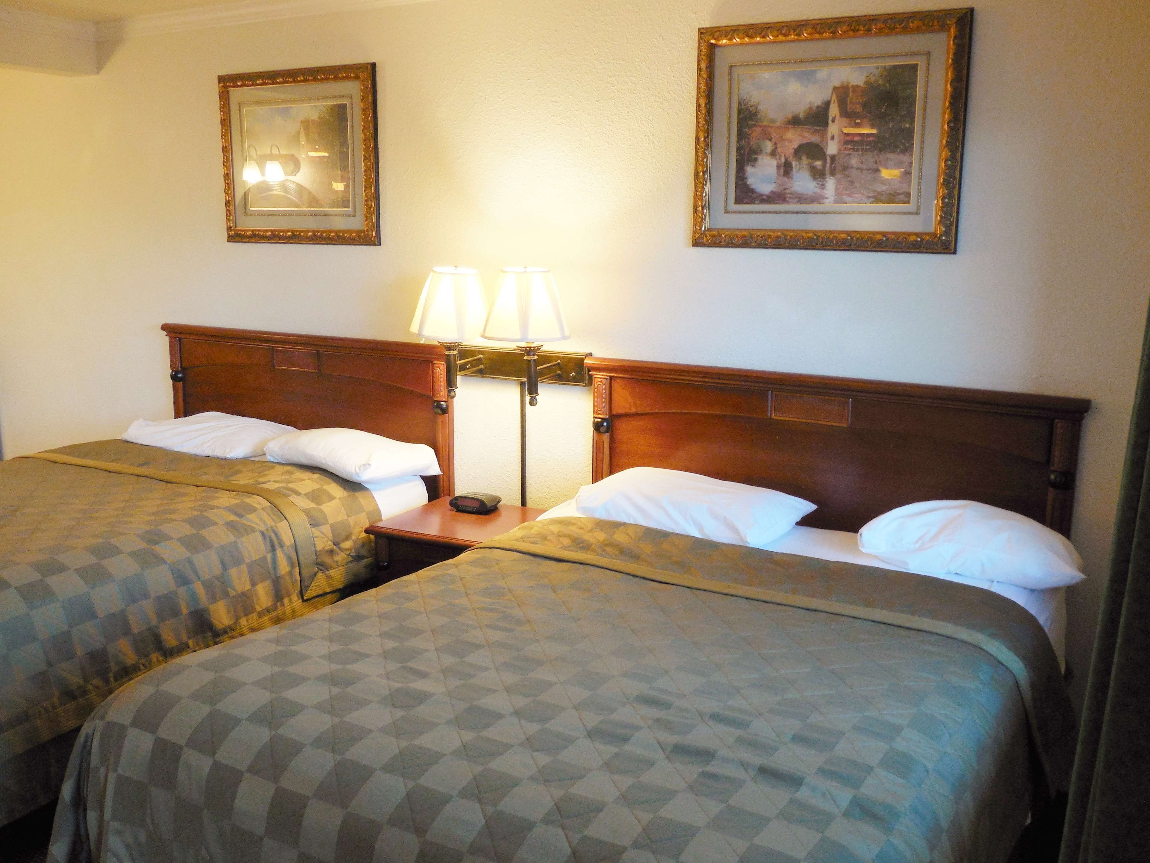 Quarto de hotel com duas camas, cada uma com cabeceira de madeira, mesa de cabeceira com abajur e duas molduras na parede.