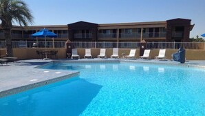 A swimming pool with lounge chairs and umbrellas, a palm tree, and a building with balconies in the background. - Americas Best Value Inn Hanford (Hanford)