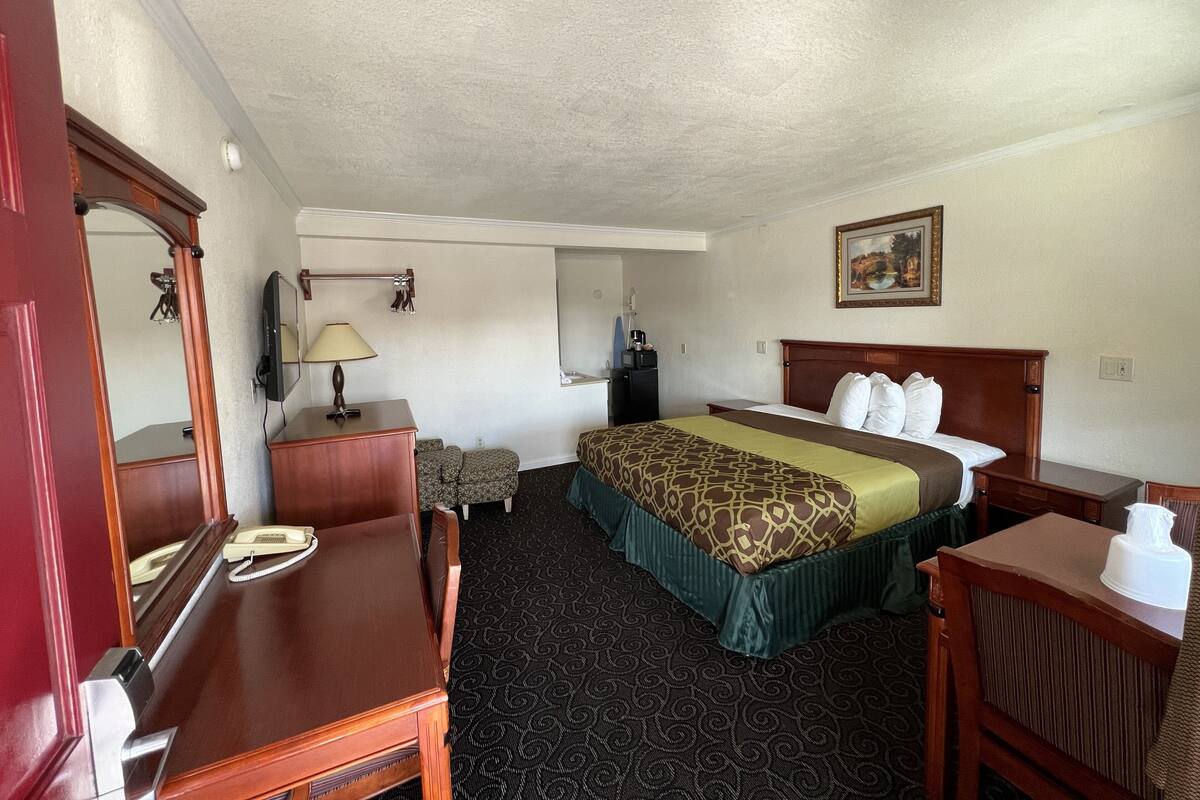 A hotel room with a bed, a desk, a chair, a dresser, a mirror, a lamp, a small table, and a painting on the wall.