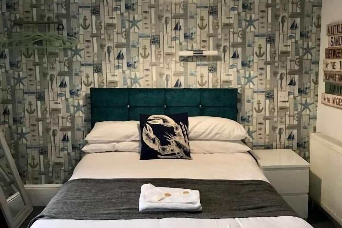 A neatly made bed with a teal headboard, a pillow with a nautical design, and a bedside table with a lamp. The room has a patterned wallpaper with nautical elements.