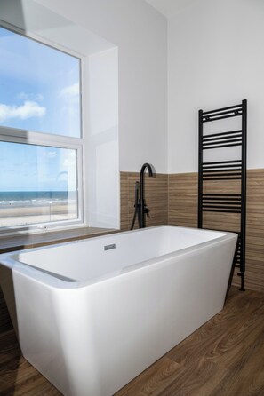 A modern bathroom with a freestanding bathtub, a window with a beach view, and a wooden wall panel. - The Craig-Y-Don Hotel (Blackpool)