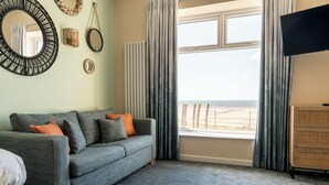 A living room with a grey sofa, orange cushions, a flat-screen TV, a large window with curtains, and a view of the beach. - The Craig-Y-Don Hotel (Blackpool)