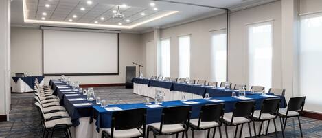Una sala de conferencias con una larga mesa, sillas, una pantalla de proyector y piso alfombrado.