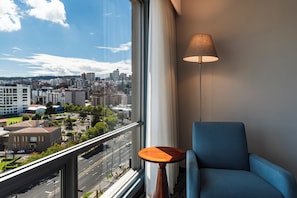 A room with a view of a cityscape through a large window, a blue armchair, a small wooden table, and a floor lamp.