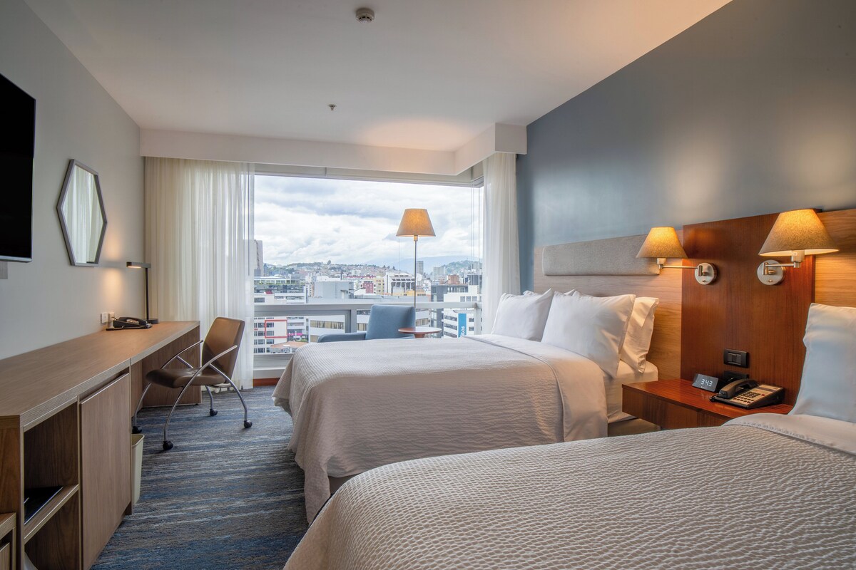 Habitación de hotel con dos camas, un escritorio, una silla, un teléfono y vistas a la ciudad.