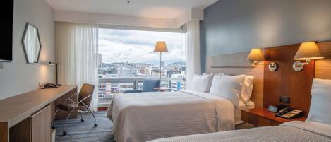 A hotel room with two beds, a desk, a chair, a phone, and a view of the city.