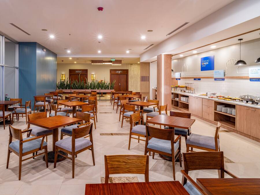 Un comedor amplio con varias mesas y sillas de madera, una barra de buffet con diversos alimentos y un ventanal con vistas a la ciudad.
