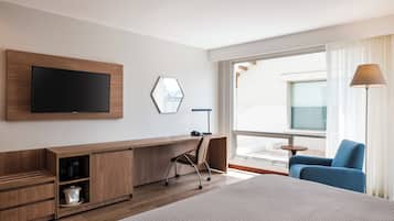 A hotel room with a bed, a desk with a chair, a television, and a large window with curtains.