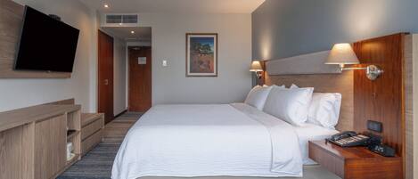 A hotel room with a large bed, a desk with a phone and a clock, a TV mounted on the wall, and a painting on the wall.