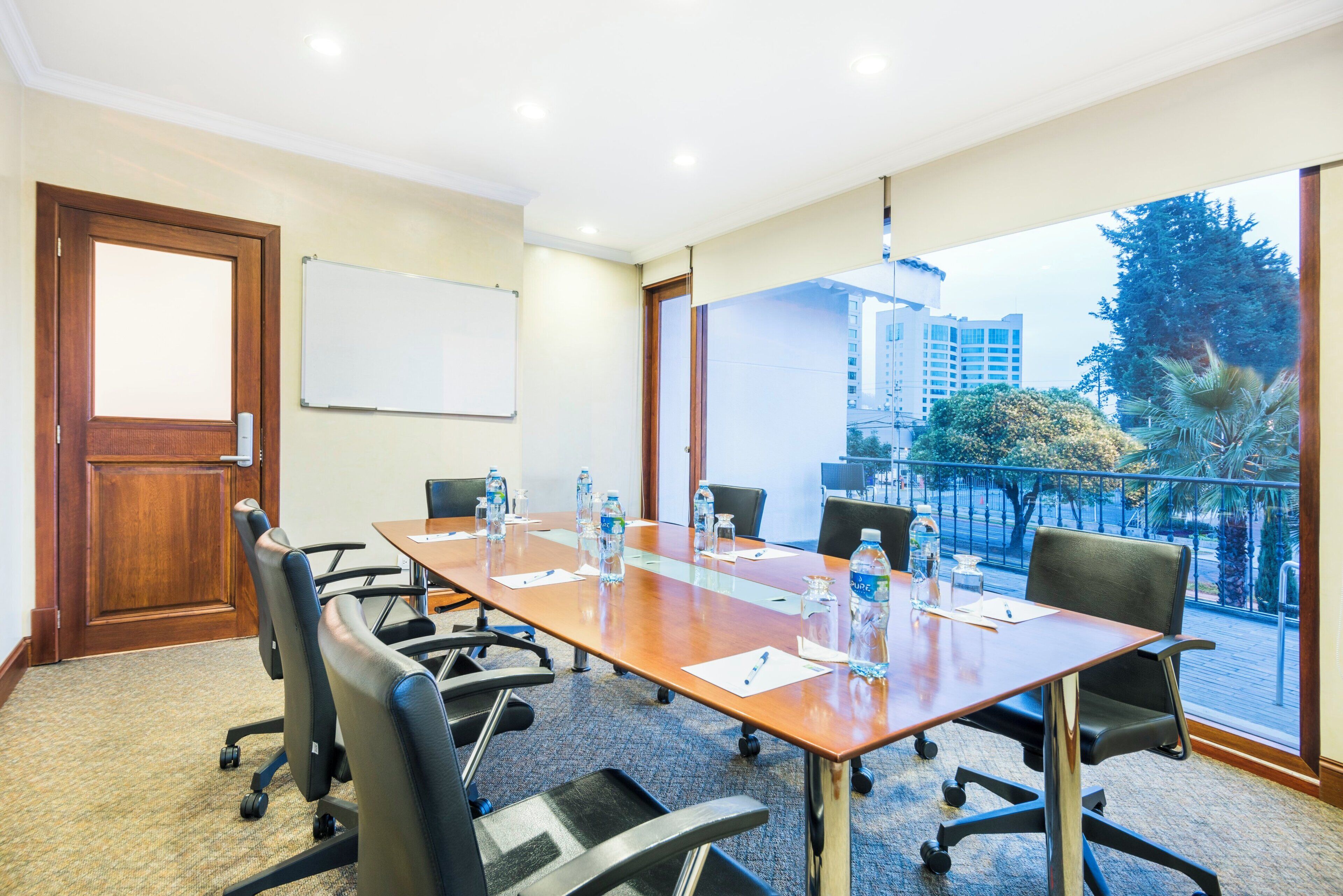 A conference room with a long table, chairs, a whiteboard, and a view of buildings and trees through large windows.