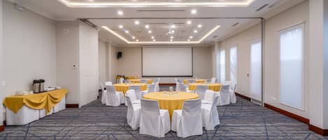 Un salón de conferencias con varias mesas redondas, sillas blancas y una pantalla para proyector.