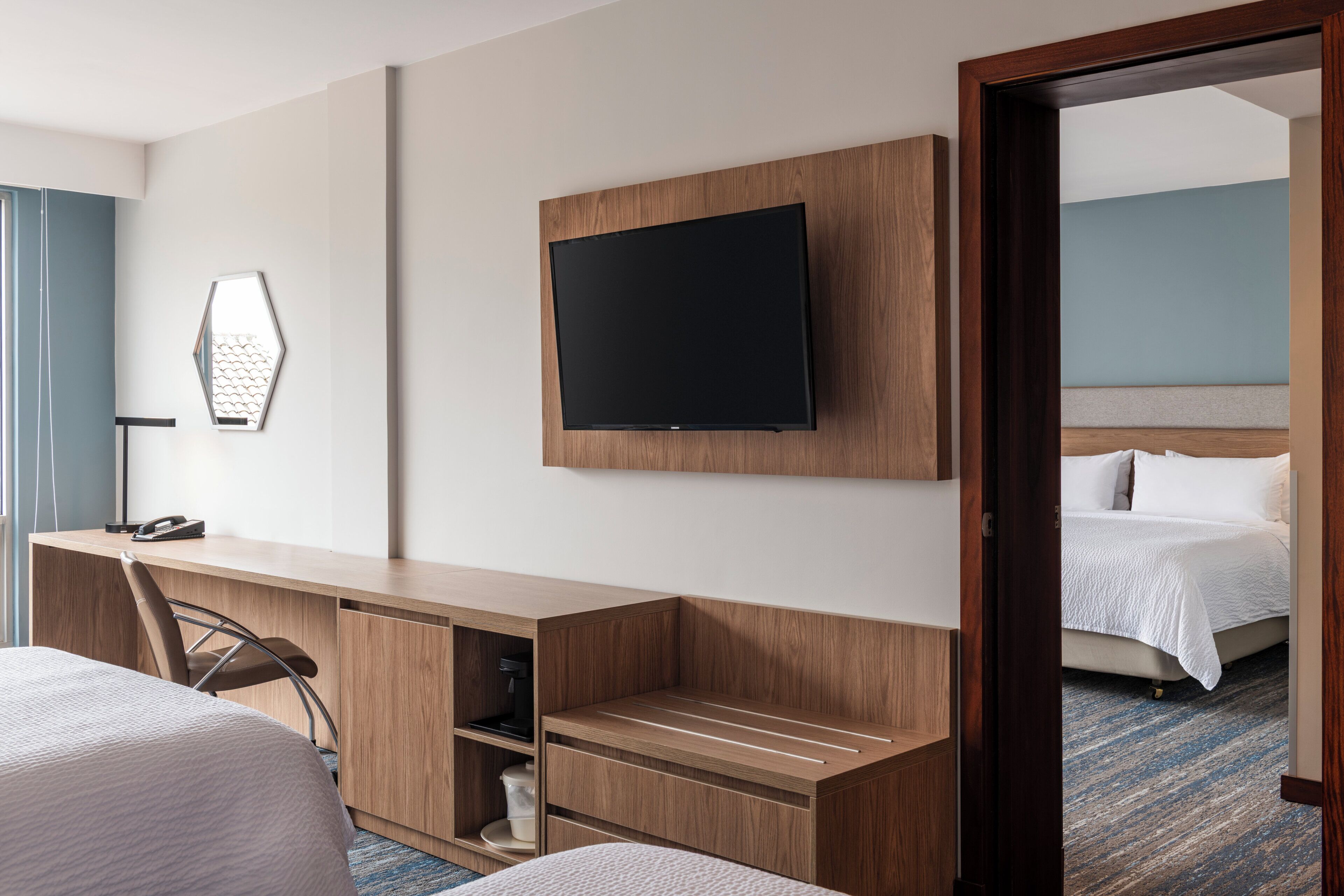 A hotel room with a bed, a desk with a chair, a television mounted on the wall, and a door leading to another room.