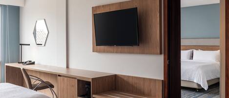 A hotel room with a bed, a desk with a chair, a television mounted on the wall, and a door leading to another room.