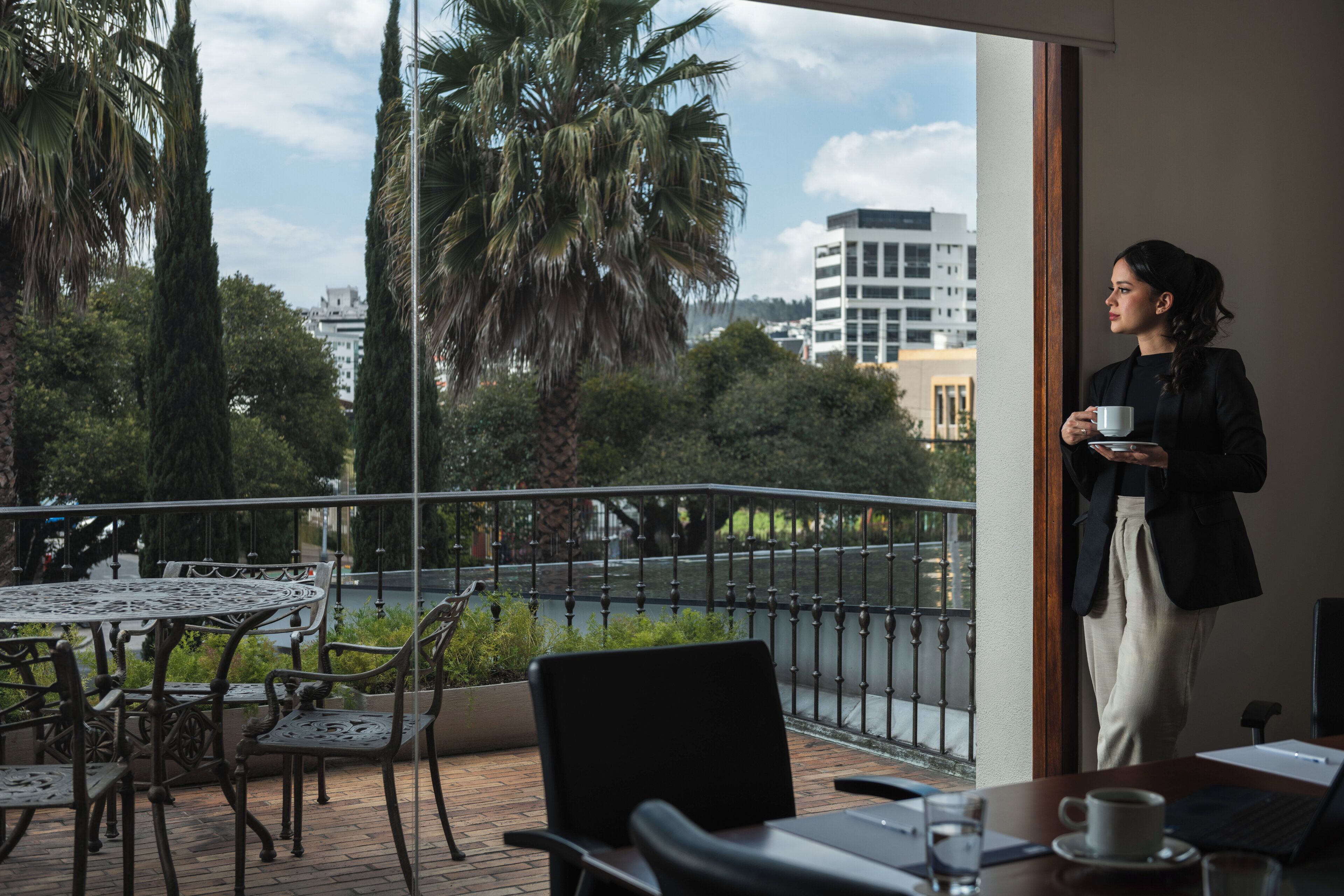 A balcony with a table and chairs, a view of trees and buildings, and a person holding a cup.