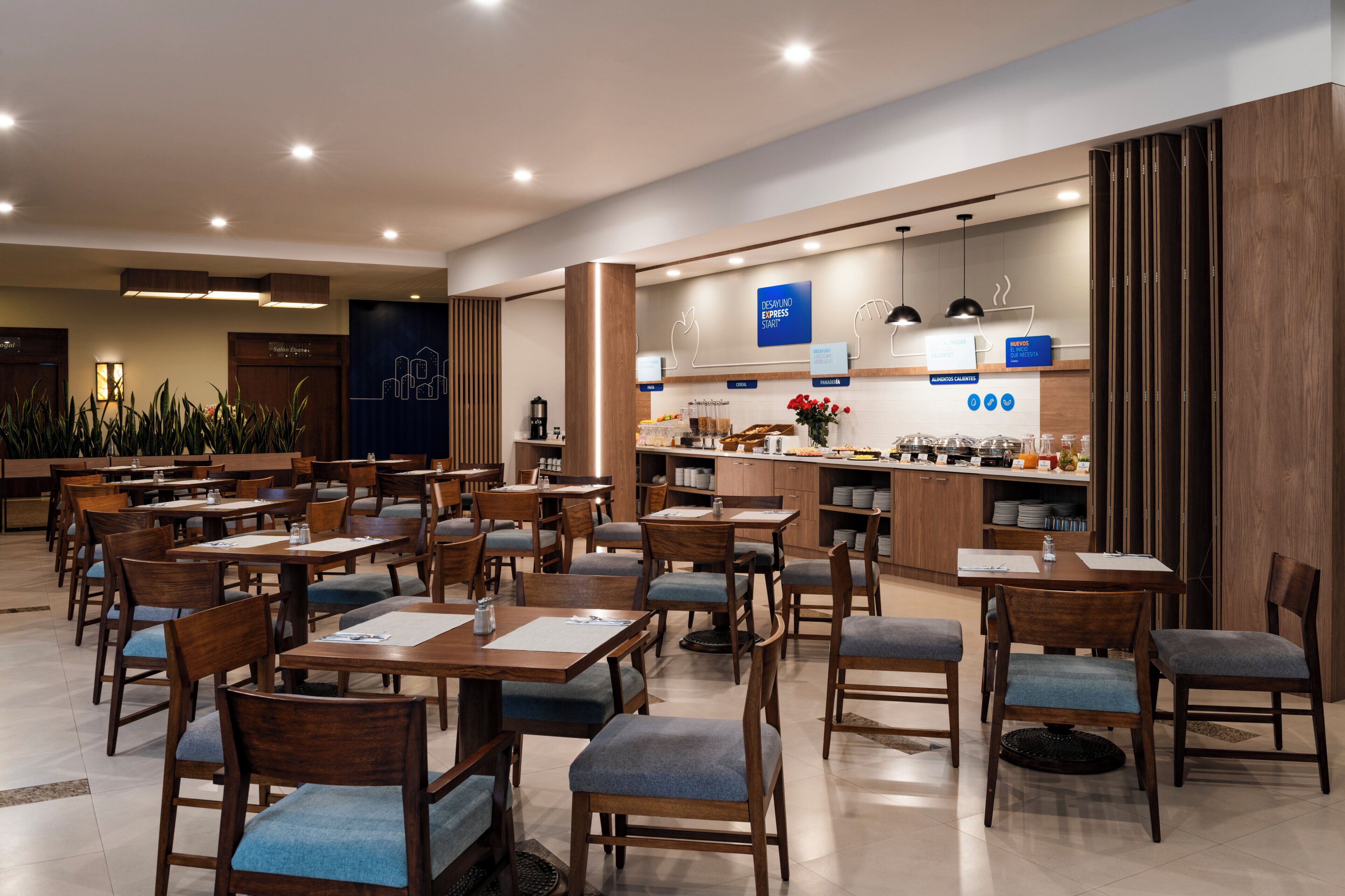 A modern dining area with wooden tables and chairs, a kitchen counter, and a sign indicating a 'Desenvolimento Express Start' area.
