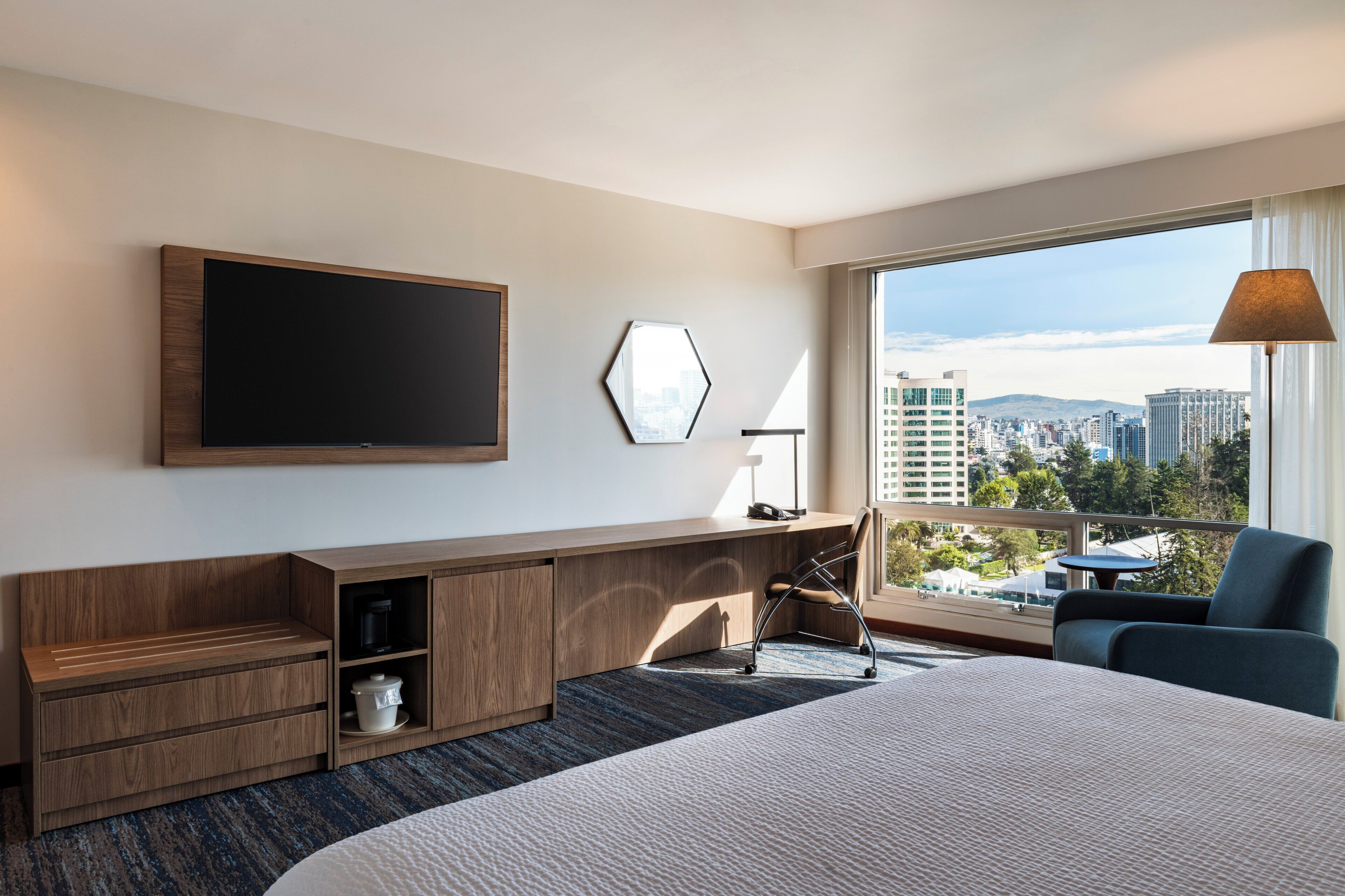 A hotel room with a large bed, a flat-screen TV, a desk with a lamp, and a view of the city.