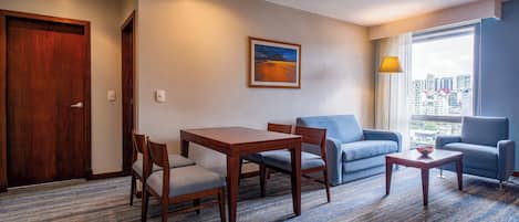 Habitación de hotel con una mesa de comedor de madera, dos sillones azules, un sofá, una pequeña mesa de centro y vistas a la ciudad por la ventana.