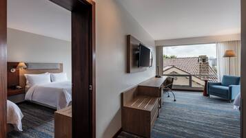 Habitación de hotel con cama, escritorio, silla, televisor y balcón con vistas.
