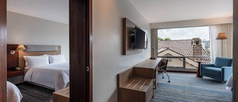 Habitación de hotel con cama, escritorio, silla, televisor y balcón con vistas.