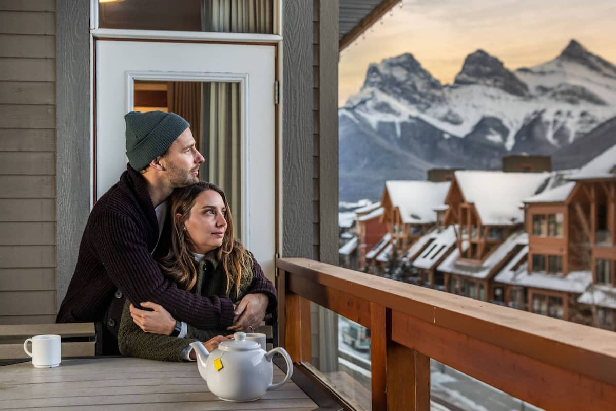 A couple sitting on a wooden deck with a view of snow-covered mountains and traditional wooden buildings.