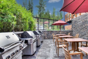 Une terrasse extérieure dotée de plusieurs barbecues, de tables en bois et de parasols rouges.